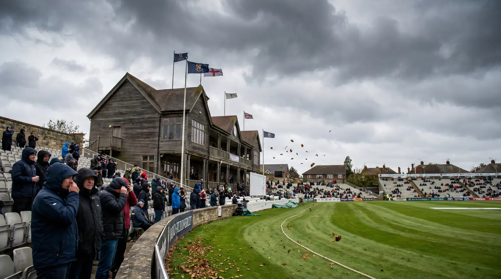 Exposed cricket ground on a windy day with flags blowing showing conditions that suppress scoring rates